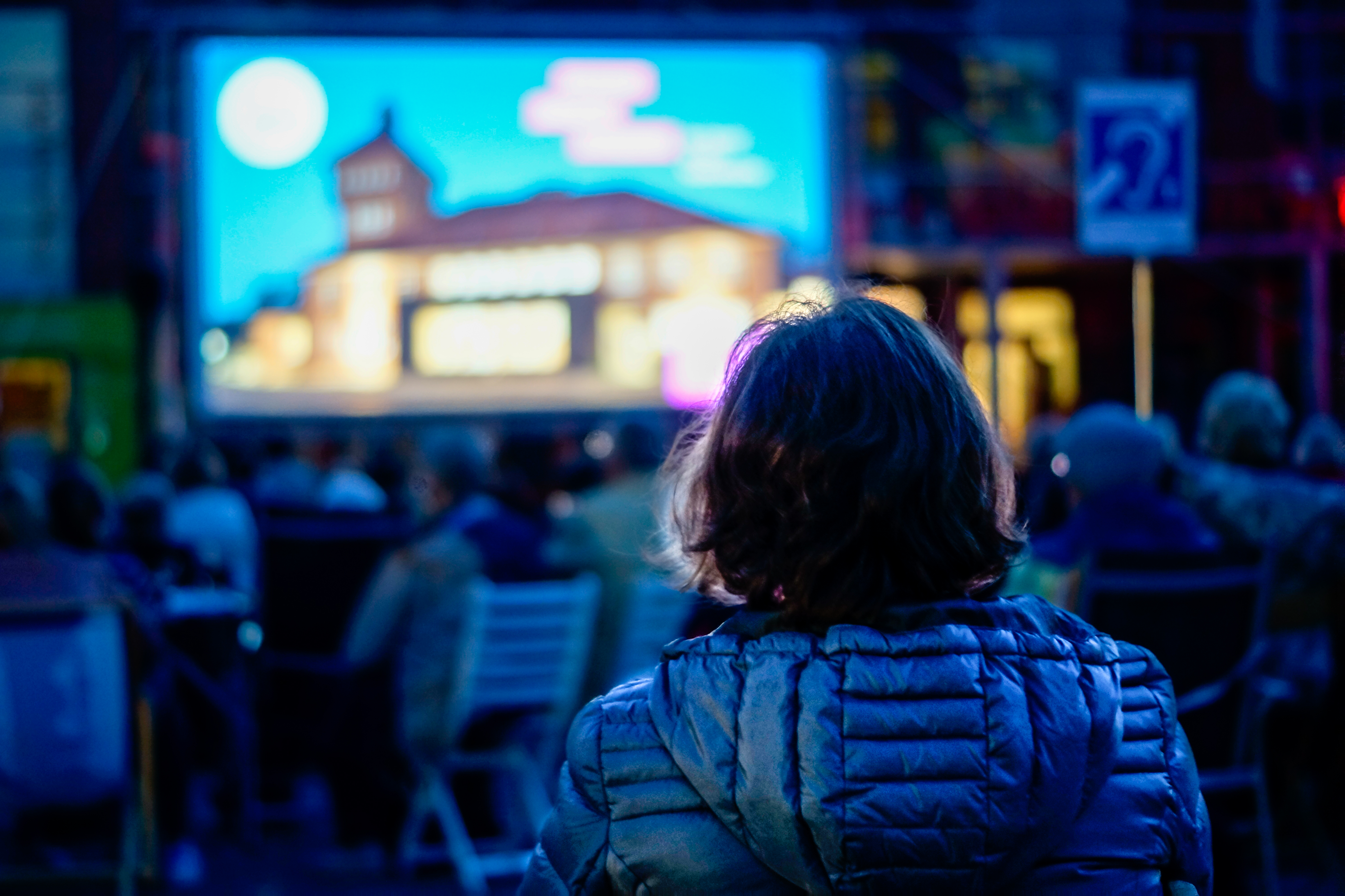 Eine Person in einer Jacke sitzt im Freien vor einer großen Leinwand, umgeben von anderen Zuschauern. Die Leinwand zeigt ein unscharfes Bild.